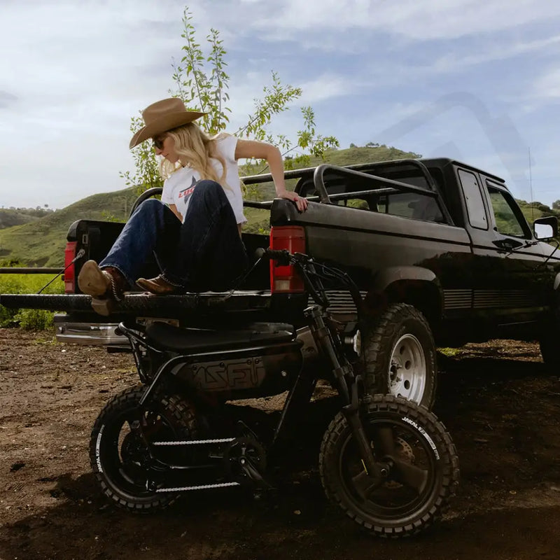 A woman wearing jeans, a white t-shirt, and a brown cowboy hat is sitting on the tailgate of a pickup truck parked in front of a green hill. A SUPER73-MZFT is parked on the dirt in front of her. @color_blackout