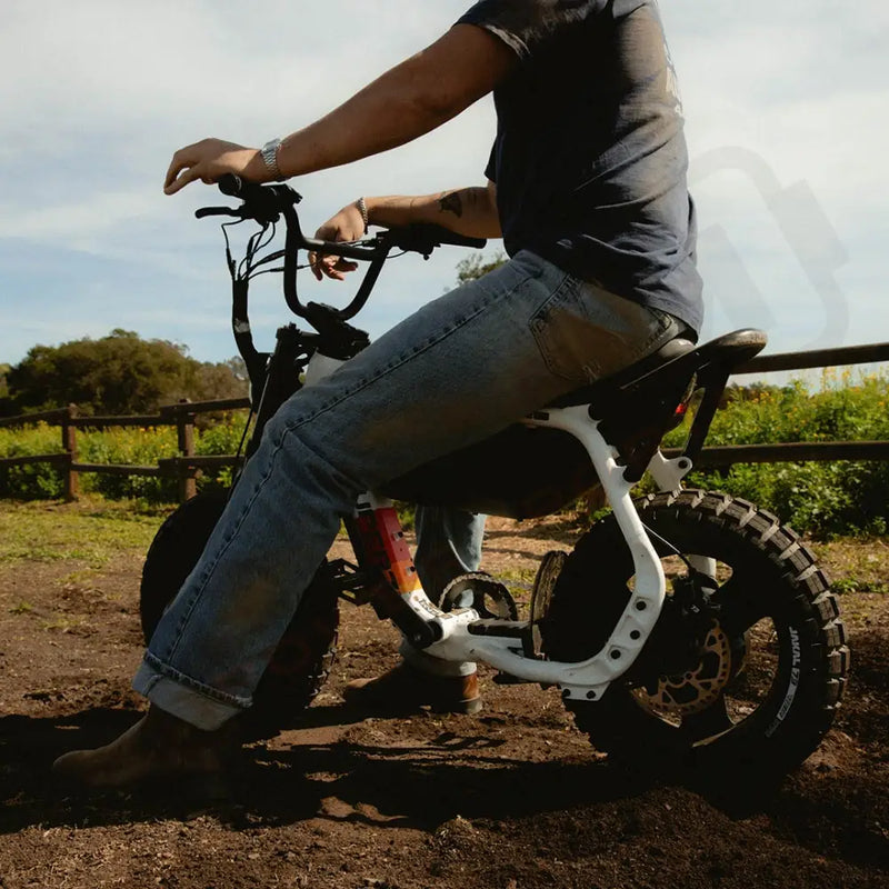 A man wearing jeans and a t-shirt, who is slightly cropped out at the top, is sitting on a SUPER73-MZFT SE ebike on a dirt path. A green field and wooden fence are visible in the background. @color_baja