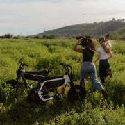 Two women, both in jeans and t-shirts, are standing in a large green field with their backs to the camera. Two SUPER-73 MZFT SE ebikes are parked in the field, just to the left of the women. @color_baja