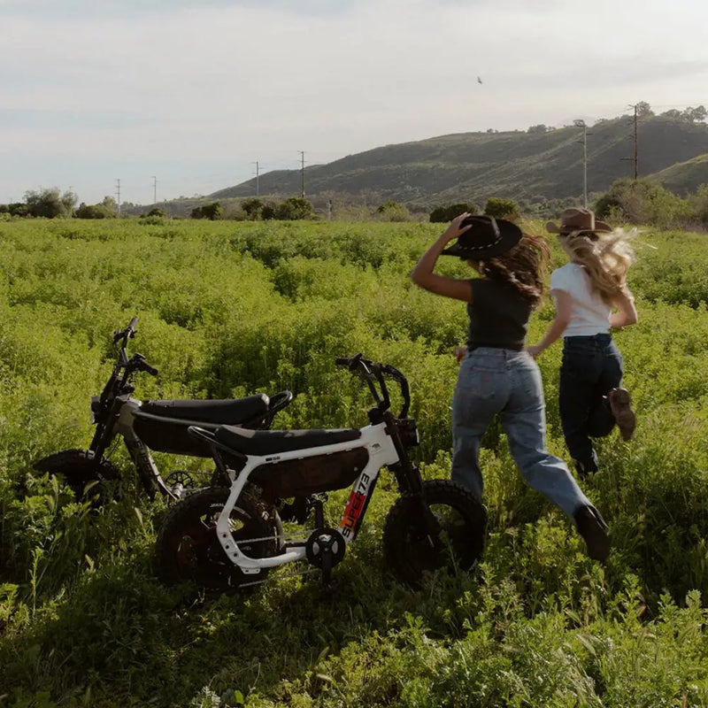 Two women, both in jeans and t-shirts, are standing in a large green field with their backs to the camera. Two SUPER-73 MZFT SE ebikes are parked in the field, just to the left of the women. @color_baja