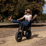 A woman in jeans, a white t-shirt, and a cowboy hat is riding a SUPER73-MZFT SE along a dirt path with a large tree and wooden fence in the background. She is kicking both of her feet up and smiling. @color_blackout