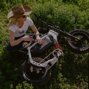 A woman wearing dark jeans, a white t-shirt, and a brown cowboy hat is sitting cross-legged on the ground in a green field. A SUPER73-MZFT SE ebike is laid on its side in front of her and she has one hand on the frame of the bike. @color_baja