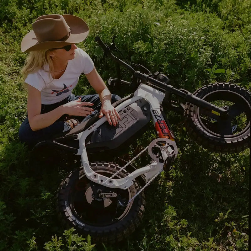 A woman wearing dark jeans, a white t-shirt, and a brown cowboy hat is sitting cross-legged on the ground in a green field. A SUPER73-MZFT SE ebike is laid on its side in front of her and she has one hand on the frame of the bike. @color_baja