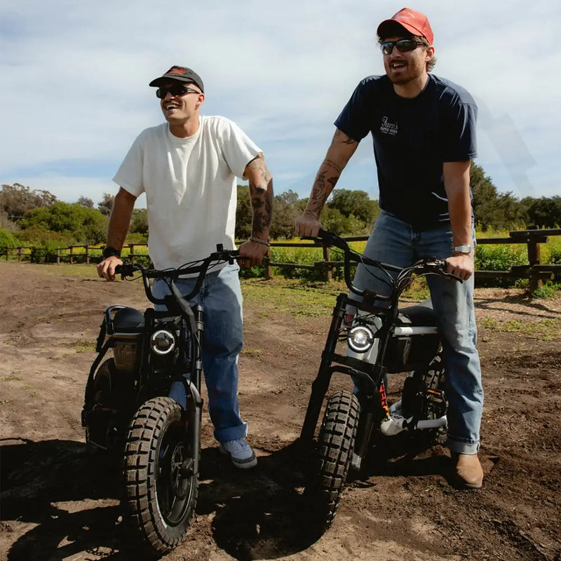 Two men, both in jeans and t-shirts, standing on a dirt path with green fields and a wooden fence in the background. The man on the left is standing next to a SUPER73-MZFT SE ebike in Blackout and the man on the right is straddling a SUPER73-MZFT SE ebike in Baja. @color_baja