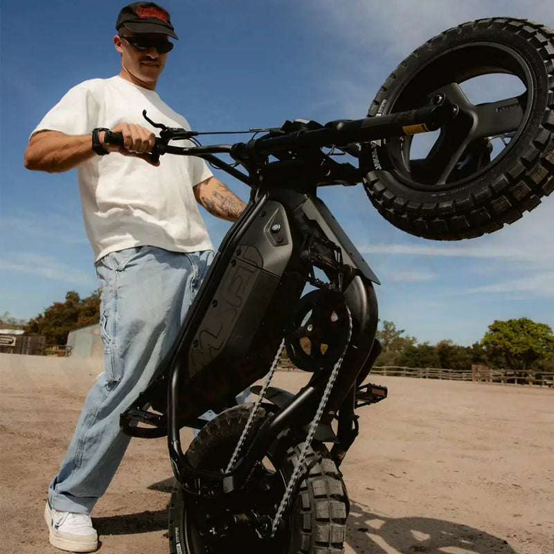 A man wearing jeans and a white t-shirt standing on a dirt path and holding a SUPER73-MZFT SE in a wheelie position. @color_blackout