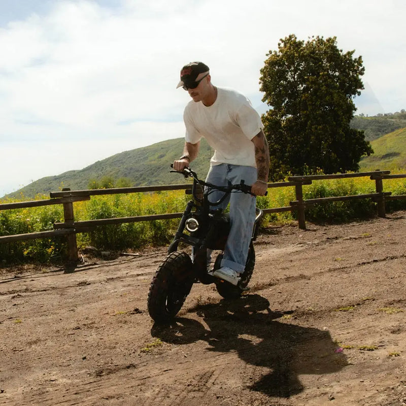 A man in jeans and a white t-shirt riding a SUPER73-MZFT SE on a dirt path with green hills in the background. @color_blackout